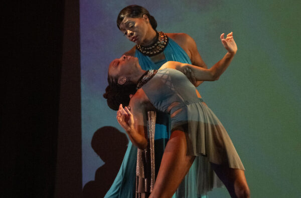 Two women dance together onstage in front of a green and purple background